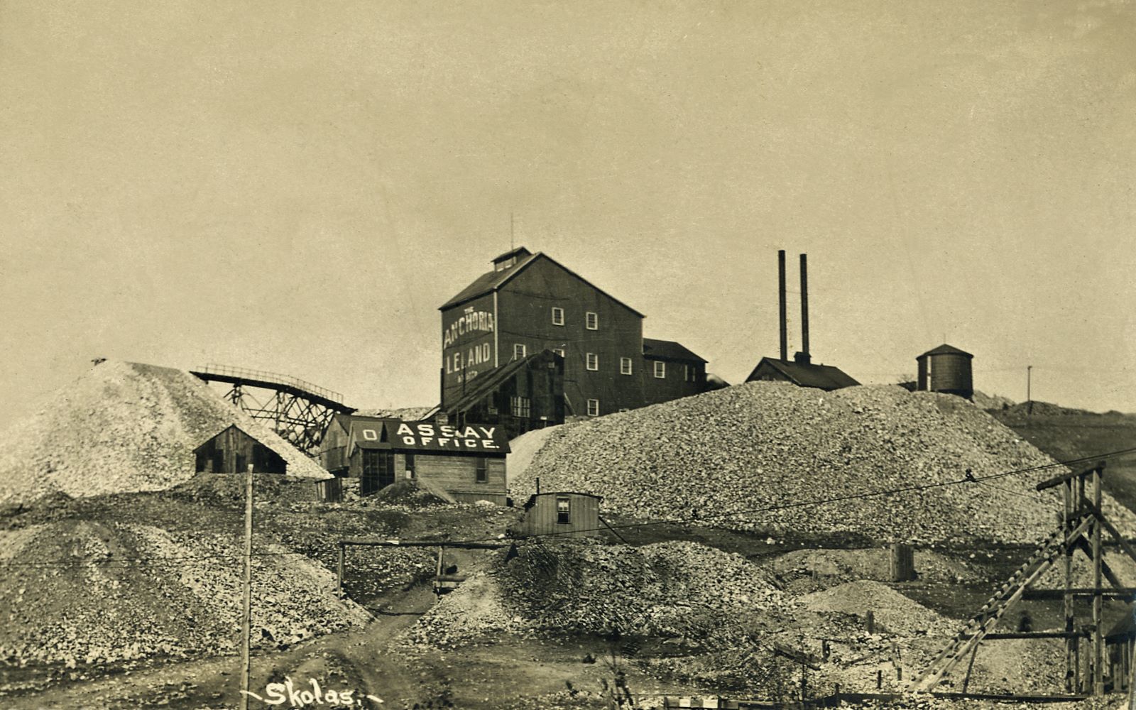 This view towards the Anchoria Leland Mine on Gold Hill is a good view of the side of the Shaft House, and while my card had some damages to it, it was simple to paint them out and create the view presented here. It is a good view to tell the look of this mine from southwest of it, down near the location of the loop on the original High Line.
While this card is marked as a Julia Skolas photo, I saw on my 1200dpi scan that there is some text that is scratched out in the dump part below the small cabin about 1/5 up from bottom. I can't read it though, the job was very well done, but it either hold just a text info about this scene, or possible another name as I've seen it happen before, and I am a little unsure if this is really a Julia Skolas photo or if she only got the rights for it. I wonder this due to the fact I can see the Trolley hanging wire in foreground right, and this part of the loop on the original High Line that was below the Anchoria Leland mine was abandoned in 1903, but Julia Skolas did not arrive in the District before about 1906-1908. The sources various a little, most say 1907, one says 1906/1907 and one say 1908. Hence, I wonder a little if she took this herself or bought the rights from whoever took it a few years earlier…