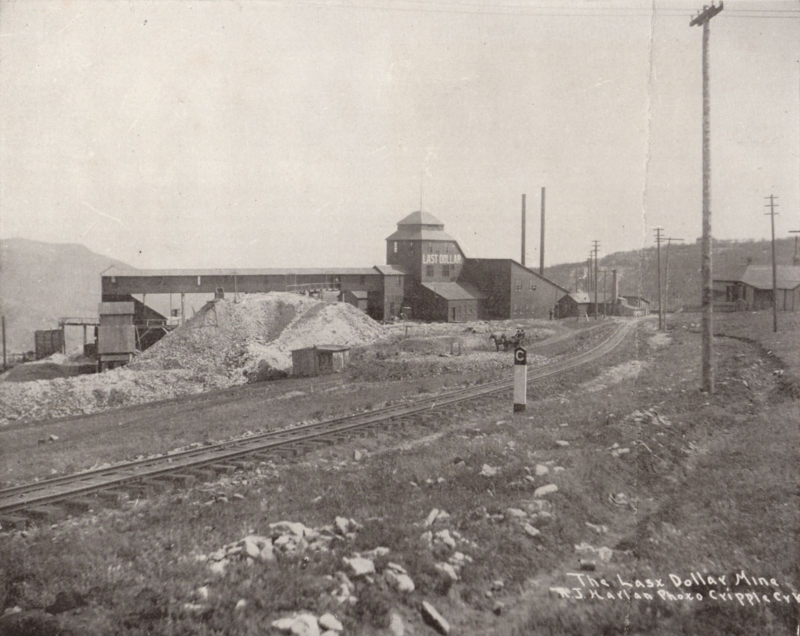 This view of the Last Dollar Mine is photographed same day and time as a more common seen view from nearly same location, where the photographer Harlan as moved closer to the Golden Circle trackage seen near lower left corner, to avoid that telegraph pole in the foreground near right-hand side.
In both views, there is a man dumping an ore-car about 1/4 in from left-hand side on this view, in front of the covered gangway from mine to the ore-house seen near left-hand side. The ore-house was connected to another Golden Circle spur, which also went on to other mines around the town of Independence, while the track in the foreground is the mainline going towards Victor mine high up on Bull Cliffs/Hills.