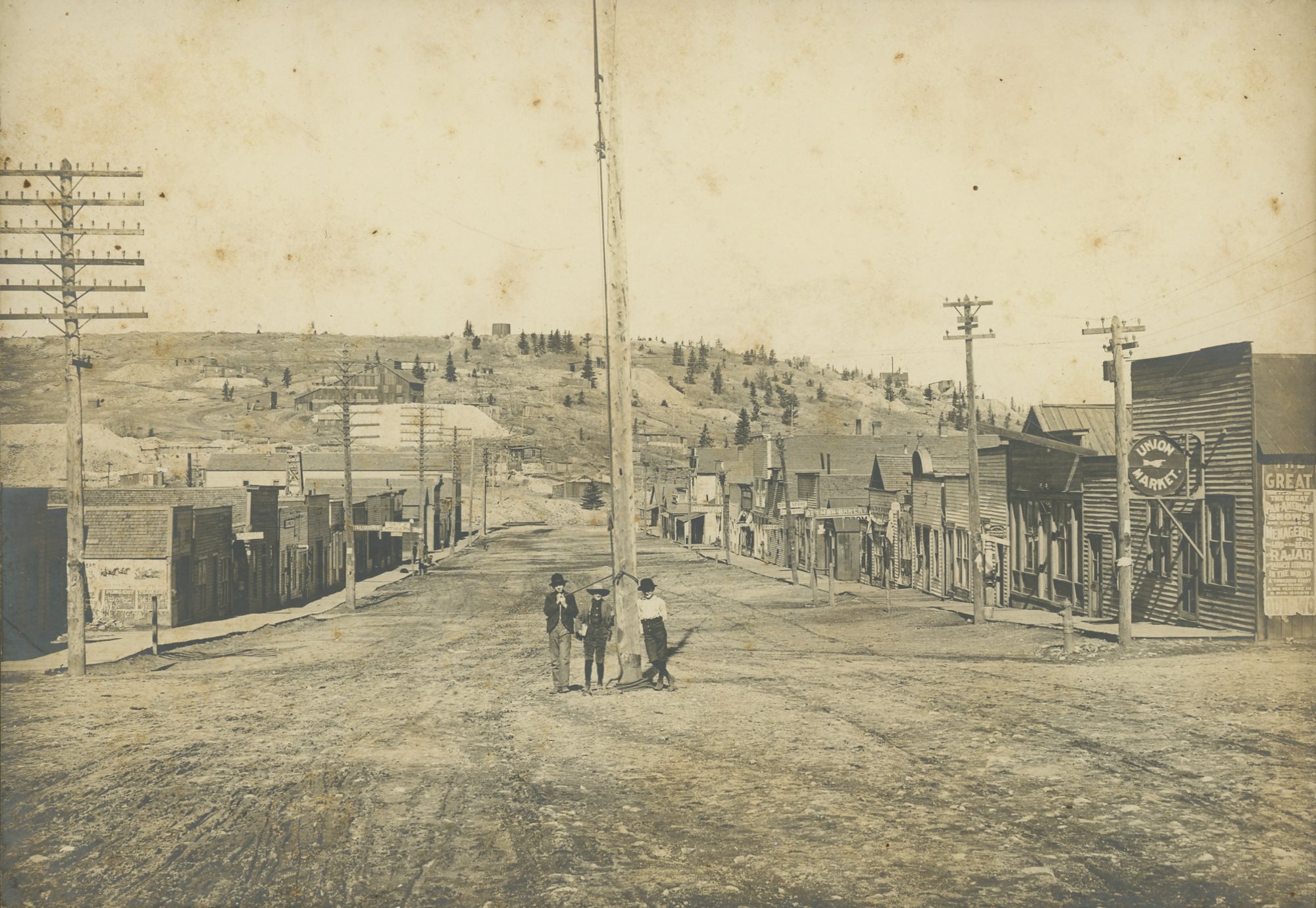 Street view from Altman, Colorado, looking west. Three boys are posing at the town Flagpole, top of Bull Hill is in the background and in the foreground at the right-hand side, we see a street sign marking the Union Market store. Further down the same side of street there are more signs, reading; Altman Bakery, Drug Store—times two. Left side of the street there are signs for; City Meat Market, Restaurant, Assaying. There is also an Assay office structure at the far end of the street, behind the small three just left of the flagpole.
   In the background some mine structures can be seen, the one large in left side is the Zenobia Mine with a dump extending out in front and right of the shaft house. The one just to the left of the flag pole I am not sure off as there are to many claims crossing that ground and I have not seen that structure before marked in any way, and further to the right, more into the image I am also at a loss at to what mines the structures are linked to as there are so many claims crisscrossing this hill.