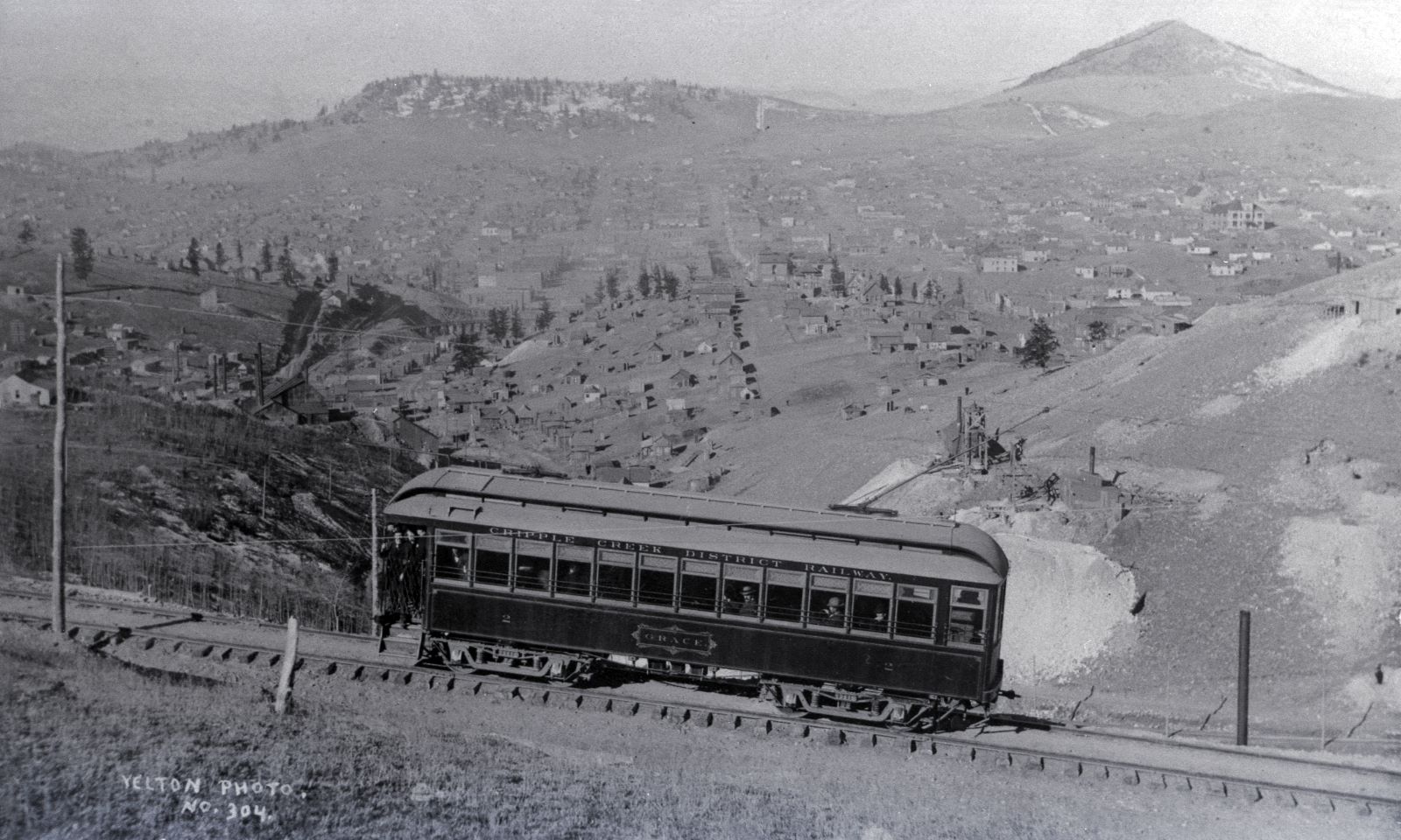View is a negative type of Photo taken by a camera of an older type of Photograph, with not the best quality film so it is rather grainy sadly. But, the view is nice, of the Trolley car named Grace climbing Gold Hill high above Poverty Gulch, along the original steep High Line grade.
* The Smokestack poking up near lower right-hand corner behind the Trolley is part of the Chicago Tunnel operation, while the two Head-frame type of mine operations seen just above the lower end/back end of the Trolley is as far as I can tell operations on the May Queen lode claim on the left and the Granite Hill lode claim on the right.
* About middle top/down and about 1/4 in from left-hand side is the structures of the Abe Lincoln Mine, and way down in the gulch one see the M.T. Wye Trestles and to the left of them is the spur to the top of the Midland Sampler seen cutting up the hill from a spur a little down from the Abe Lincoln.