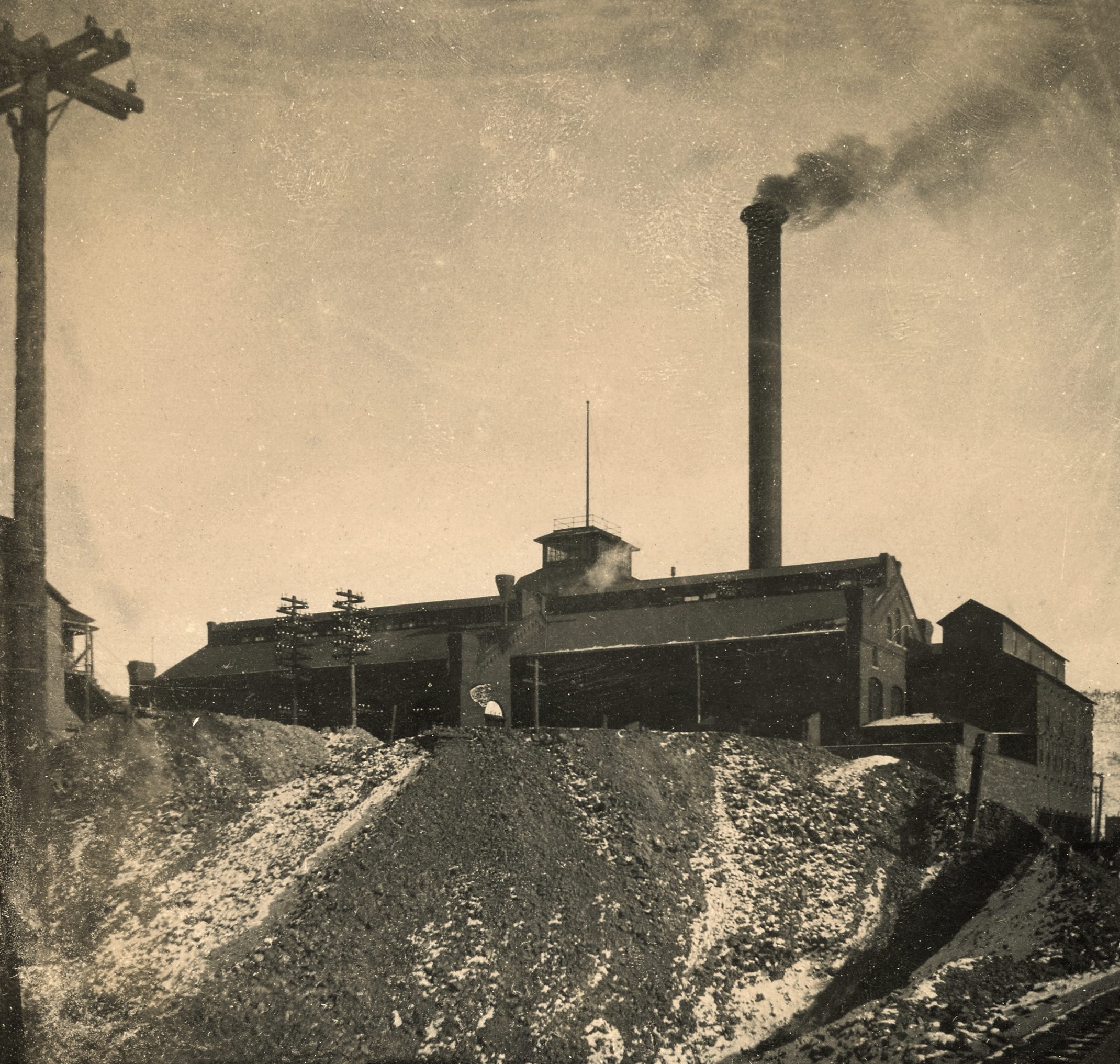 This view of the Gold Coin Mine is taken from the roadbed of the F. & C.C. railroad, as seen in lower right corner where a piece of the track is visible. While the sheet this image was pasted into has an 1898 date to it, that is plainly wrong as this is the brick version of the mine, and that only happen after it burned in 1899, so I think this is more like 1900, but I can't date it more precisely, sadly.
   The version shown here is enhanced to fix some fading issues on the original, in addition to me using a perspective filter to make the sides of the structures more appealing and level, making me having to crop the image a little of non-important pieces, and I also had to paint in some extra sky and track pieces, but I think it gives the credit to the source image as it should do, a view of the mine as it would have been seen from the Florence & Cripple Creek tracks west of the mine!
   I did procure the colored version of this image, if that is what you see. Source was gray-toned, or in common speech black & white. Used an online service and tweaked and worked with image to get what looks best to my eyes for the moment.