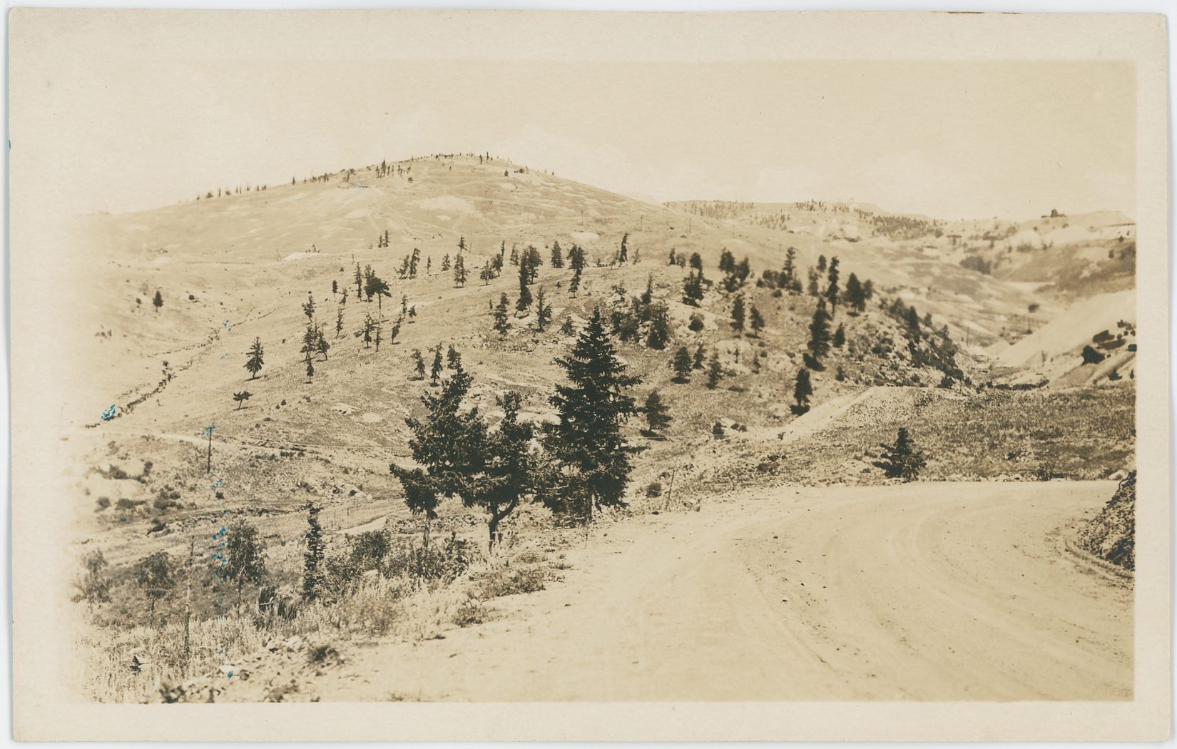 I am pretty certain this is on the former main road between Victor (behind and to the right) & Cripple Creek (into image towards left) where it went over the hills and valleys at a lower elevation then the railroads. I further believe this is as the road turn down into Eclipse Gulch, and that the valley up on the right-hand side is the Eclipse Gulch.
* The dump seen about 1/3 down from the top sticking into the view from right-hand edge would be the huge dump left after the Economic Mill burned down, at least that is what I think the dump is.
* Near the same edge, against the sky, the Blue Bird Mine can be seen as a darker spot, while the American Eagle is almost faded into the sky further to the left, about 1/4 in from right-hand side.
* The Shaft House of the old Bostwick Mine can be seen about 1/3 down from top and about 2/7 in from left-hand side.