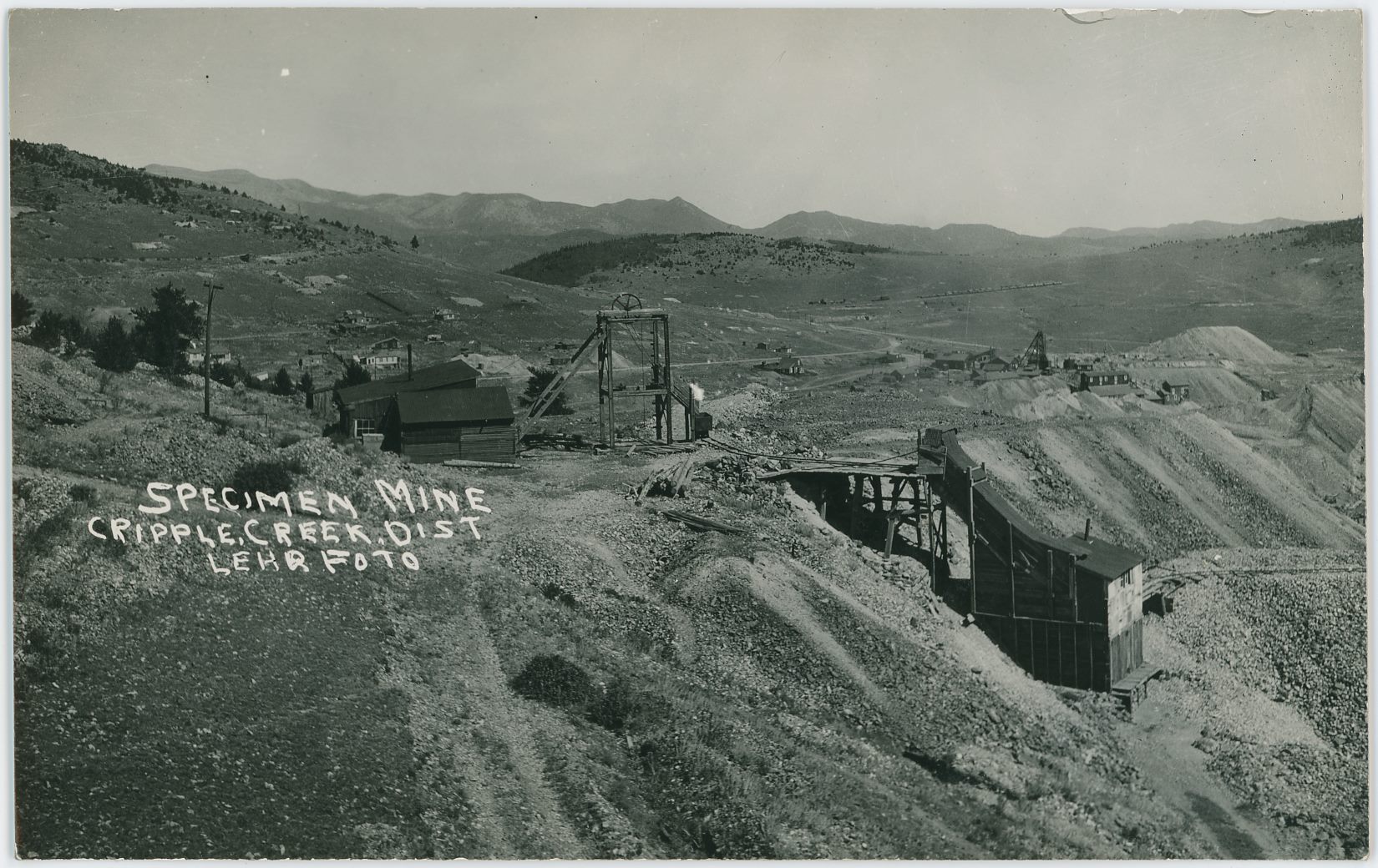 Direction of this view is east, northeast, on Bull Hill, just west of the surface structures of the Specimen Mine operation, with an open Head-Frame and Hoist-house and shed linked to it, and a small Ore-house to the right down the hillside a small bit.
   In the distance, up from the orehouse, the surface structures of the Vindicator No. 1 Mine are seen with a large Head-frame and Ore-house down into massive dumps, impossible to tell if it still is linked to a railroad or not in this view. Further on in the distance to the left, a big string of railroad cars say that the Midland Terminal is still in operation, but there is no trace of the High Line Electric Division, as where that should be I only see a road surface – cutting almost the headframe of the Specimen Mine in two so to speak.
   Image is not sharp enough to tell if there are any rails left on the old Golden Circle tracks seen climbing Bull Hill, or is it Bull Cliff, about 1/4 down from top and about 1/4 in from left-hand side, but I do think the standard gauge M.T. tracks are there still when this photo was taken by Lehr many years ago.