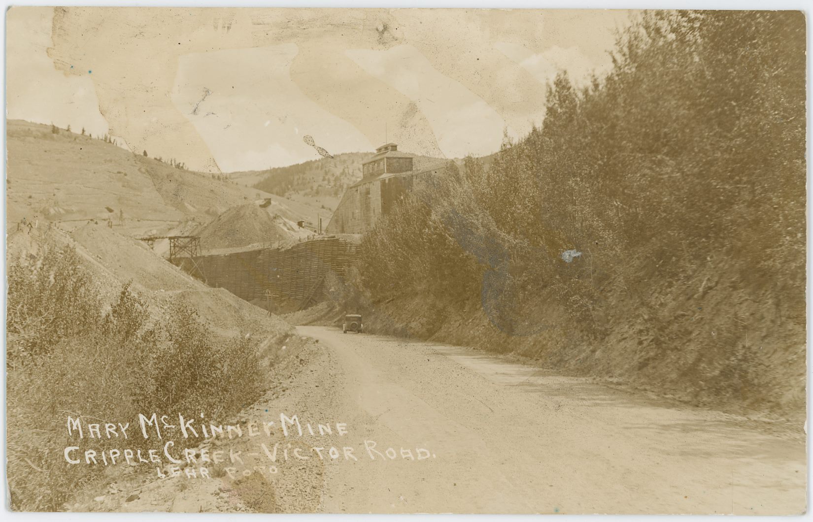 This view is from along the Victor-Cripple Creek road, running on the former roadbed of the Florence & Cripple Creek, looking towards the Mary McKinney Mine about center sideways and its huge crib-wall. If one could have walked into the view and hitched a ride with the automobile seen one would have ended up in Cripple Creek.
   The Hill in distance left-hand side is part of Gold Hill, and I wonder if part of the old Short Line roadbed is visible up there near the skyline. I can't sadly see anything though in a high-resolution scan of 1200dpi, as there is just too much blur, and overall the whole card is sadly not in a good shape due to someone at some time in the past having spilled something over it.